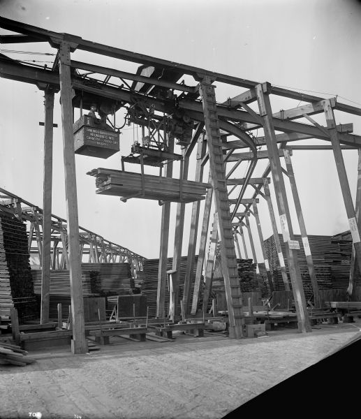 Pawling & Harnischfeger 3-ton monorail hoist with lumber handling unit in the Pacific Lumber Co. lumberyard. There's a man in the cab and the text stamped on the side of the cab reads "Pawling & Harnischfeger Builders, Milwaukee, Wis., Capacity 6,000 Lbs., No. 2291." In the background is a sign that says "Smoking Prohibited."