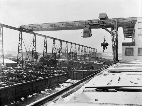 Pawling & Harnischfeger semi-gantry crane with a magnetic hoist unit in a foundry yard. The crane is stamped with the text "Pawling & Harnischfeger Builders, Milwaukee, Wis." The crane is suspended above a railroad car and large stacks of cord wood with a crane runaway. A large industrial tank is in the background.