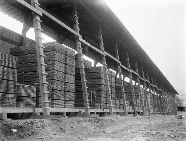 A Pawling & Harnischfeger 3-ton monorail hoist in lumber storage racks. There's a man in the cab, and another man standing at the bottom of one of the stacks of lumber. The cab is stamped "Capacity 6,000 Lbs., No. 2702."