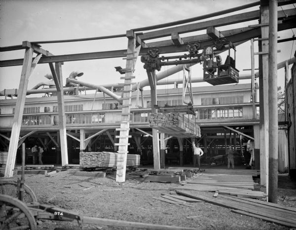 Pawling & Harnischfeger 3-ton monorail hoist crane with lumber unit lowering its load at American Lumberman. There is an operator in the cab, and men on the ground guiding the load onto a pallet. The cab is stamped with the test "Pawling & Harnischfeger, Milwaukee, Wis., Capacity 6,000 Lbs., No. 2156."