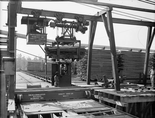 Pawling & Harnischfeger 3-ton monorail hoist crane with a special 5-ton floor transfer car in a lumber mill. There is an operator in the hoist, and there are two men on the ground operating the floor transfer. The monorail hoist is stamped "Pawling & Harnischfeger, Milwaukee, Wis., Capacity 6,000 Lbs., No. 2299" and the floor transfer is stamped "Pawling & Harnischfeger Builders, Milwaukee, Wis., Capacity 30,000 Lbs., No. 2373."