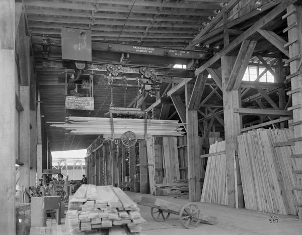 A standard Pawling & Harnischfeger 3-ton monorail hoist with lumber handling unit in a lumber mill. There is an operator in the cab, as well as a man with a hat on the ground in the background of the picture.