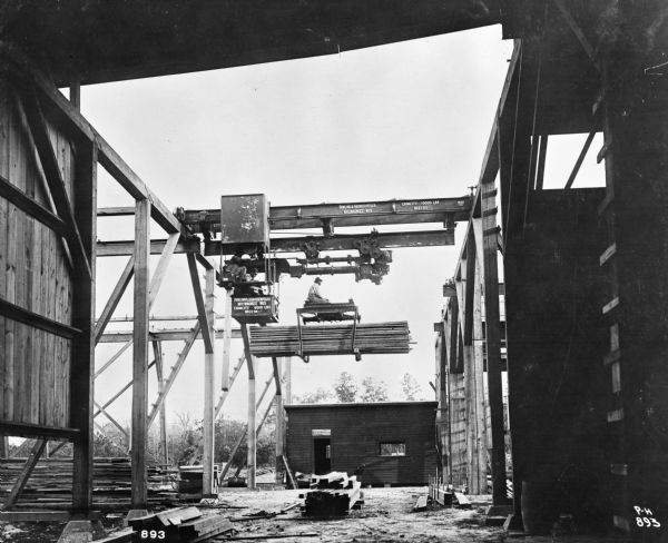 Pawling & Harnischfeger 3-ton monorail hoist with lumber handling unit operated by a man. There is a small shed in the background. The I-beam is stamped "Pawling & Harnischfeger, Milwaukee, Wis., Capacity 6000 Lbs., Patented, Pat. No. [illegible]" and the cab is stamped "Pawling & Harnischfeger Builders, Milwaukee, Wis., Capacity 6,000 Lbs., No. 2194."