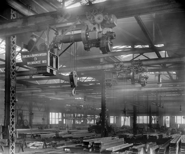 Pawling & Harnischfeger 5-ton type "M" auxiliary hoist at the Belmont Iron Works. A man wearing a hat is in the cab. The monorail hoist crane, used in steel fabrication, has a pendant cage and dial controls. It is stamped with the text "Pawling & Harnischfeger Co., Milwaukee, Wis." and "3110." The crane in the background is stamped "Pawling & Harnischfeger, Milwaukee, Wis., Cap. 10,000 Lbs., Cr. No. 3113, Pat No. [illegible]."