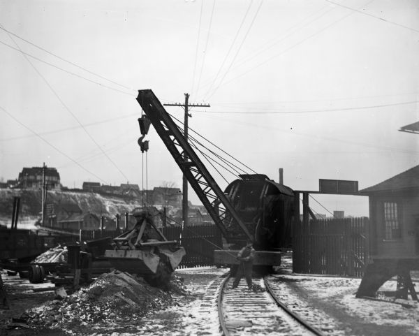 Pawling & Harnischfeger gasoline powered crane mounted on a 5-ton standard flatbed railroad car with a grab bucket. A man is standing on the ground on railroad tracks in front of the railroad car. He is holding a rope attached to the bucket. There are houses on a hill in the background.
