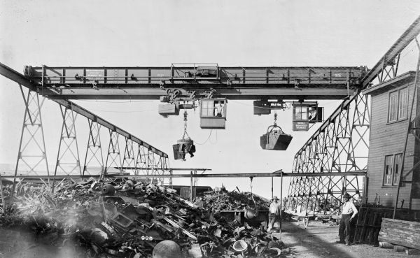 Pawling & Harnischfeger 3-ton transfer crane with two 3-ton monorail hoists. The hoist on the left has an electromagnetic unit, and the one on the right has a grab bucket attachment. The transfer crane is stamped "Pawling & Harnischfeger Builders, Milwaukee, Wis., Capacity 6,000 Lbs., No. 1930, Patented, Patent Nos. [illegible]" The monorail hoist cab on the left is stamped "Pawling & Harnischfeger, Milwaukee." There are men in the cabs and men on the ground looking at the camera near a large pile of scrap metal. The location is Simmons Co.