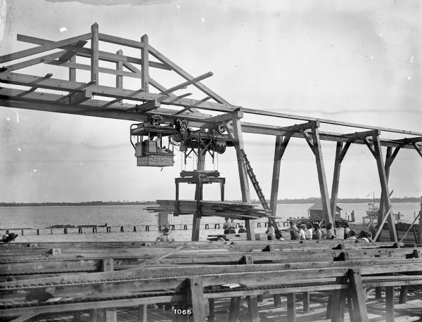 Pawling & Harnischfeger 5-ton monorail hoist with a lumber handling unit preparing to load lumber onto a boat in what is possibly Pensacola, Florida. Two men are in the cab, and a group of men are standing on the ground. There is a pier and boat near water in the background. The crane is stamped "Pawling & Harnischfeger Co., Milwaukee, Wis., Capacity 10,000 Lbs., No. 3255," the lumber unit is stamped "U.S. Patent No. 982387[?], Mexican, 11769, Canadian, 133513[?], Others Pending. Pawling & Harnischfeger" Co., Milwaukee, Wis."