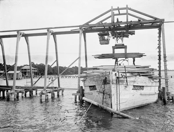Pawling & Harnischfeger 5-ton monorail hoist with a lumber handling unit, possibly in Pensacola, Florida. The monorail track extends over the dock to permit loading barges and boats with lumber. The text on the front of the ship reads "Ainsley" and there's a man in the cab, men on board, and men on the dock. A man and woman stand on the end of the pier, and a Lumber Co. building with porches is on the shoreline in the background.