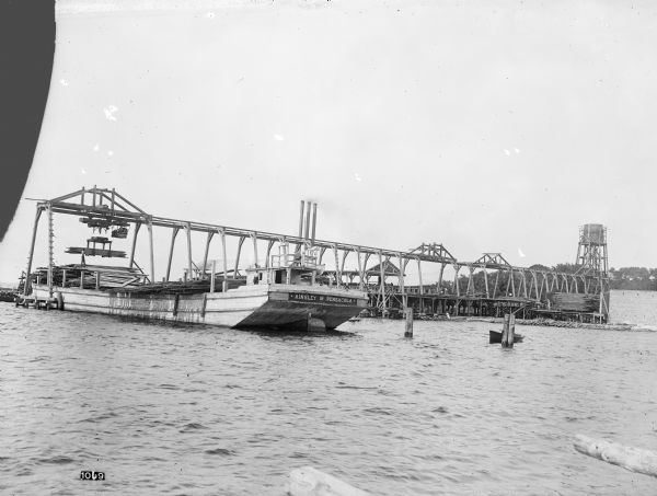 View from water of Pawling & Harnischfeger 5-ton monorail hoist with a lumber handling unit, possibly in Pensacola, Florida. The monorail track extends over and beyond the dock to permit loading barges and boats with lumber. The text on the front of the ship reads "Ainsley of Pensacola" and there's a man in the cab, men on board the barge, and men on the dock. Three chimneys can be seen in the background.