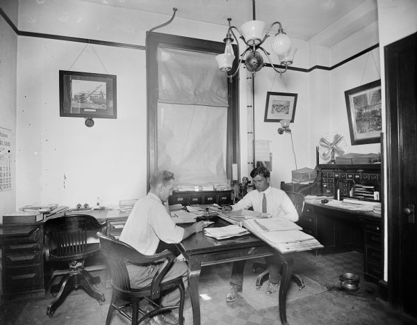 Two men working a desks in the Pawling & Harnischfeger accounting office. There are photographs on the wall, including a crane, the P&H plant, and a St. Louis Exhibition calendar. A spittoon is on the floor on the right.