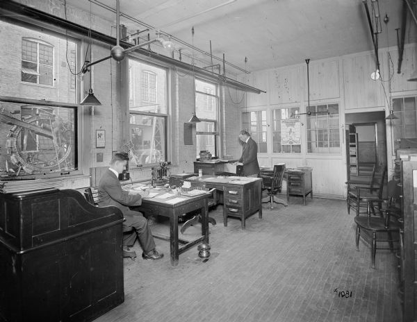Men at desks in the Pawling & Harnischfeger advertising department. Large windows on left look out towards another building, and men on equipment can be seen working outdoors.