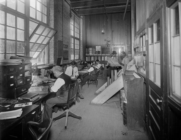 Men working at desks at the Pawling & Harnischfeger shop office. A row of windows is on the left, and another building can be seen through an open window.