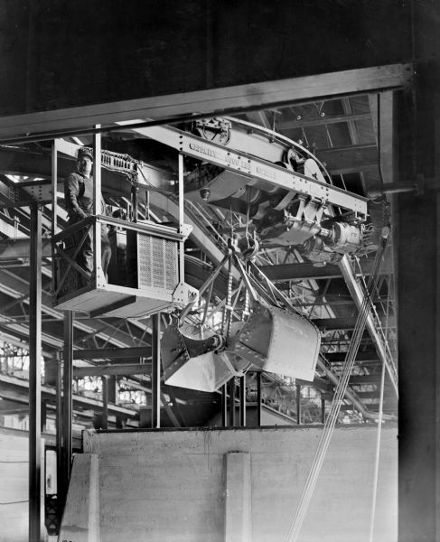 A Pawling & Harnischfeger monorail hoist with a grab bucket. The rail is stamped with the text "Capacity 6,000 Lbs., No. 3245". A man stands in the cab looking down at the camera.