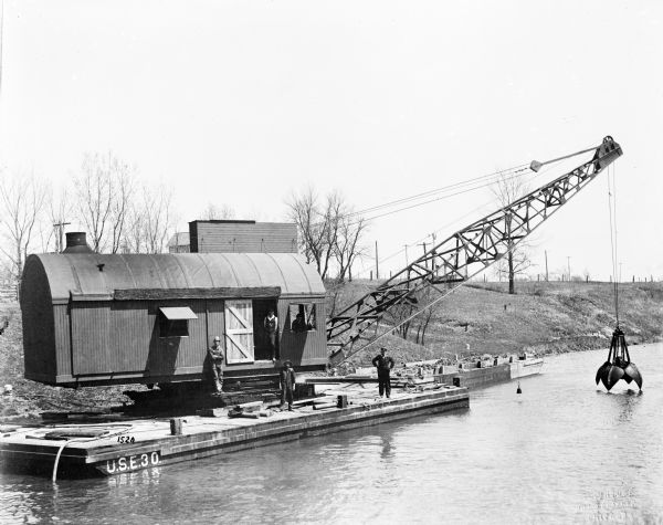 Pawling & Harnischfeger lattice crane with a dredging bucket on a gasoline powered barge. Men in work clothes and hats stand on the barge which is on a shoreline. The barge is marked "U.S.E. 30" and the lower right corner of the photograph has the text "J.L. Brouse Photographer Chicago."