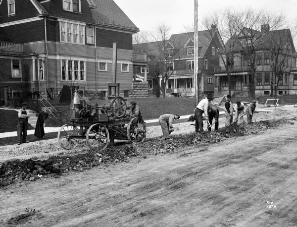 Pawling & Harnischfeger gasoline powered concrete breaker on a residential street with an operator in the cab and men working on the ground. Houses and yards are in the background.