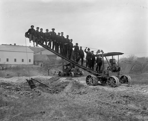 Pawling & Harnischfeger gasoline powered mobile dragline with a horizontal trench digger in the background. A large group of men are standing and posing on the deployable side discharge apparatus. Most of the men are wearing work clothes and hats. Three men on the right wear suit jackets, vests, neckties, and trousers.
