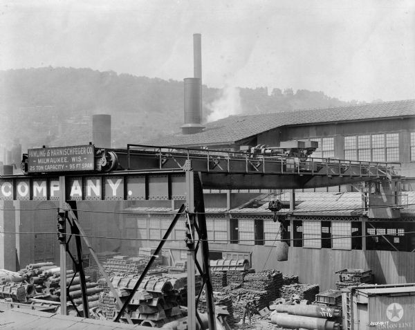 Elevated view of Pawling & Harnischfeger 25-ton type "N" bridge with a 5-ton type "A" trolley and a standard bottom block, equipped for exterior use. There is a man on the ground under the crane holding a rope attached to a hook. Another man is in the cab of the crane, which is attached to an industrial building. There are hills on a steep hill or mountainside in the background. A sign on the crane reads "Pawling & Harnischfeger Co., Milwaukee, Wis., 25 Ton Capacity, 95 Ft. Span." The date "July-19-17" is written in the lower right hand corner.