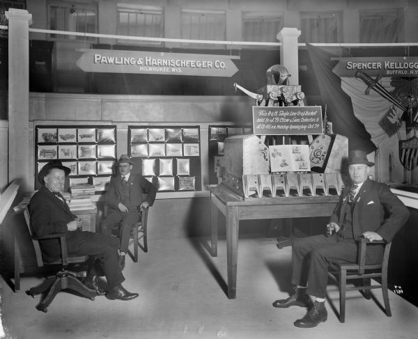 Single line grab bucket on display at P&H booth at a trade show with three men sitting around the display. The men are wearing hats and sitting in chairs, and two are holding cigars. On the wall behind them are a display of photographs. The sign above the display reads "Pawling & Harnischfeger Co., Milwaukee, Wis." The grab bucket is labeled with a sign that reads "This P&H Single Line Grab Bucket Sold to J.B. Clow & Sons, Coshocton, O. at 12:45 P.M. Monday - Opening Day - Oct 7th."