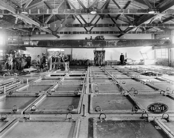 View across room of tanks and a Pawling & Harnischfeger type "GN" low overhead crane in a tanning operation. Men are standing on the left towards the back of the building near an open doorway. Hides can be seen piled behind them. Hides are hanging on a rack from the crane near a man on the right. The P&H logo is on the lower right corner and reads "Founded in 1884 By Pawling & Harnischfeger, Quality Service."