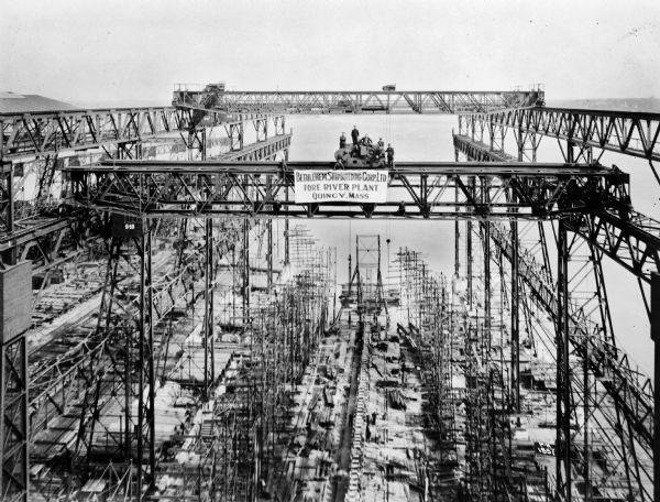 Elevated view of four Pawling & Harnischfeger lattice girder bridges trolley hoists used in shipbuilding. There are men on the bridge of the nearest crane and the banner on the crane reads "Bethlehem Shipbuilding Corp. Ltd. Fore River Plant Quincy, Mass." In the background is water and a shoreline in the distance.