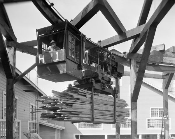 Milwaukee Electric Crane & Manufacturing Company monorail hoist with lumber handling unit on a monorail track with an operator in the cab at Long Bell Lumber Co. Another man is below the cab operator, and two buildings surround the crane.