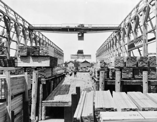 View from water of Milwaukee Electric Crane & Manufacturing Company lumber crane in an unknown lumber yard. The cab is stamped "Milwaukee Electric Crane & Mfg. Co., No. 762, 7 1/2 Tons," there is a sign that reads "The Milwaukee Crane," and there is an operator in the cab. Two men are on a pier under the cab, stacks of lumber are under both sides of the crane, and there are two buildings in the background.