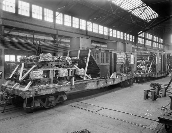 A flatbed rail car, possibly on the packing floor at the Milwaukee Electric Crane Co., loaded with trolleys, motors, cases, and other parts needed to assemble various Milwaukee Electric Crane & Manufacturing Company cranes. The sign on the crane reads "Milwaukee Electric Crane & Mfg. Co., Incorporated, Milwaukee, Wis." and "Stone & Webster, Incorporated, Engineers-Builders, Boston, New York, Chicago, San Francisco." The freight car is from the C.&N.W. Railway.
