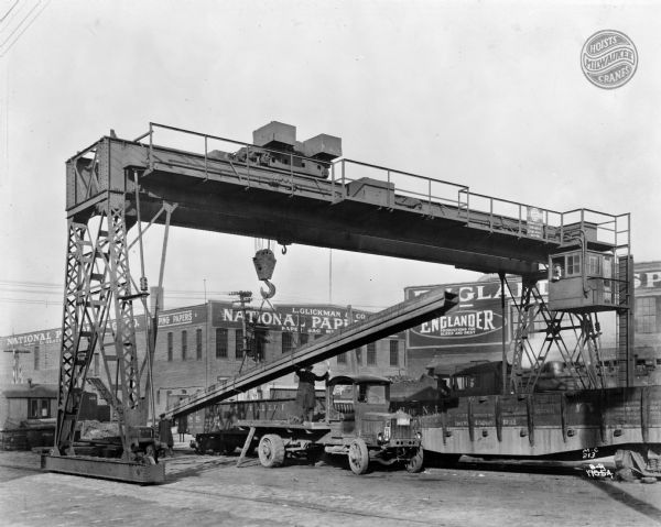 Milwaukee Electric Crane & Manufacturing Company gantry crane involved with the construction of the Long Island Railroad. There is a sign on the crane that reads "L.I.R.R. The Milwaukee Crane" and the crane is stamped "Milwaukee Electric Crane & Mfg. Co., 25 Tons, No. 442, Aux. 3 Tons." There are men helping load a truck, and the buildings in the background read "L. Glickman & Co.," "National Paper Bag Co.," and "Englander."
