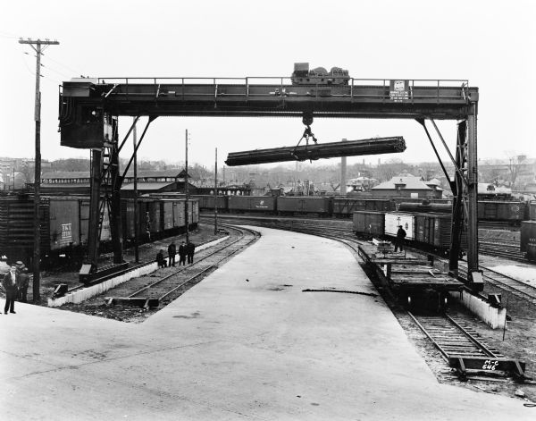 Milwaukee Electric Crane & Manufacturing Company gantry crane unloading railroad cars at the Fisher Body Co. There is a sign that reads "The Milwaukee Crane" and the crane is stamped "Milwaukee Electric Crane & Mfg. Co., 20 Tons, Aux. 5 Tons, No. 1170" and there are men standing on the ground next to the railroad tracks. In the background are industrial buildings, houses, and chimneys.