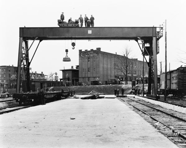 A Milwaukee Electric Crane & Manufacturing Company gantry crane with dual hooks, one standard, and the other a magnetic unit, used for unloading train cars. A sign on the crane reads "The Milwaukee Crane," and text on the building in the background reads "Central Storage Warehouse." There are men standing on top of the crane as well as on the ground.