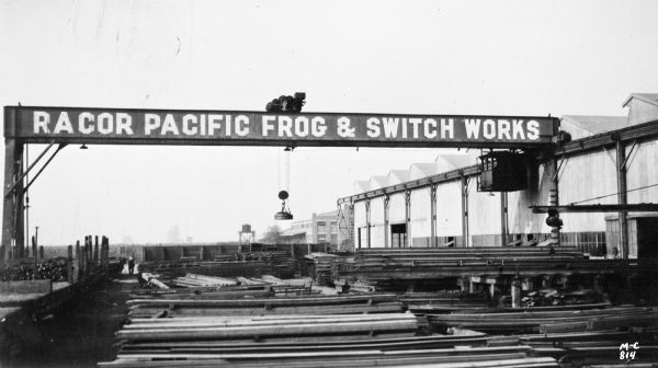 Milwaukee Electric Crane & Manufacturing Company single leg gantry crane handling steel at the Racor Pacific Frog and Switch Works. There are a couple of men working in the yard.