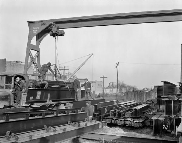 Milwaukee Electric Crane & Manufacturing Company half ton single leg gantry crane in a steel operation. There is a driver in a truck beneath the crane, and a man is standing on the bed. Two other men stand near the truck. A sign reads "Danger Keep Off." Houses and buildings are in the background.