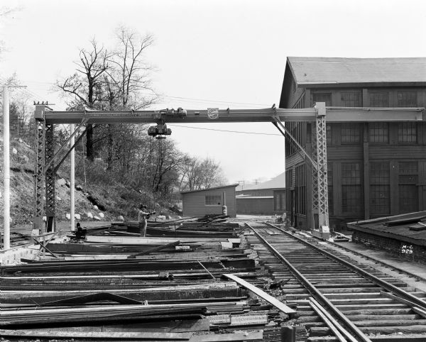 Milwaukee Electric Crane & Manufacturing Company gantry crane over a railroad track and piles of lumber, with a building on the right. There is a sign that reads "Milwaukee Electric Crane & Mfg. Co., 2 Tons, No. 1287" and there is a man on the ground.