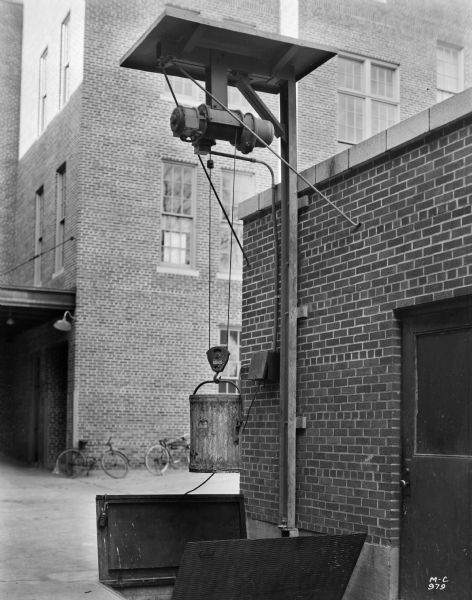 Milwaukee Electric Crane & Manufacturing Company floor operated half ton auxiliary hoist. The hoist assembly is marked "1,000 Lbs," and is above a set of delivery doors in the ground near the exterior of a building. There are bicycles leaning against a large building in the background.