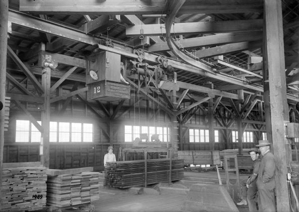 A monorail hoist with lumber carrying unit and monorail track switch, with the number 12 painted on the front, at the Port Hammond Lumber Company. Other text on the hoist reads "Pawling & Harnischfeger, Milwaukee, Wis., Capacity 6000 Lbs., No. 2294." Lumber workers wearing hats pose near stacks of lumber.