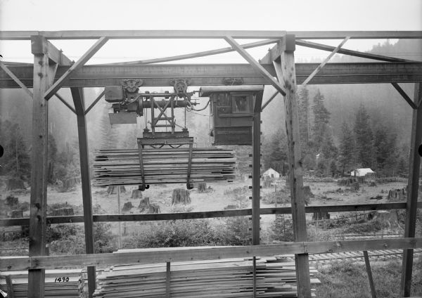 Elevated view of monorail hoist with lumber carrying unit outside at the Port Hammond Lumber Company. Tree stumps, a tent, and other small buildings are visible in the clearing in the background.