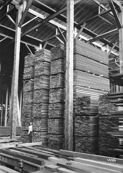 A man wearing a tie and a pocket watch on his belt, stands between stacks of lumber in a tall building at the Port Hammond Lumber Company. Tracks for the monorail hoists are visible along the beams above.