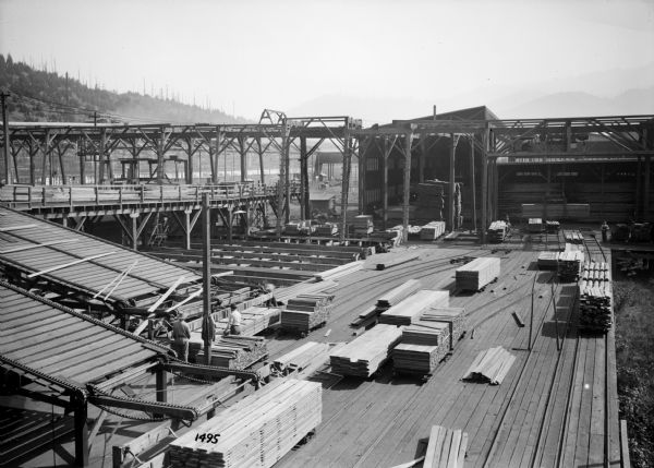 Elevated view of workings of the Port Hammond Lumber Company, including monorail hoists, tracks and men working with large stacks of lumber. There are large hills or mountains in the background.