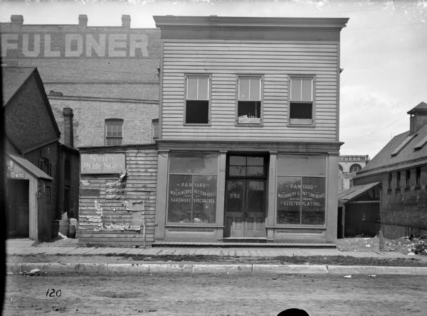 The exterior of the first Pawling & Harnischfeger storefront from across a street. The text in the windows reads "Panyard Machinery & Pattern Works," "Mfrs of Hardware Specialties," "General Jobbing, Electroplating." There are advertisements for "Swift's Pride Soap," "Drink Edelweiss," "Yeast Foam," and "Hunter Tobacco" pasted up around the entrances to the buildings. The brick building behind the storefront is painted with the word "Fuldner."