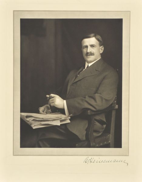 Three-quarter length seated studio portrait of Benjamin Heinemann, Wausau lumberman and banker. He has papers in his lap, and holds a pair of eyeglasses and a cigar.
