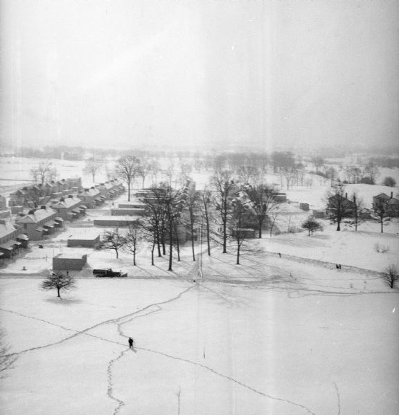 Elevated winter scene of Greendale (Milwaukee Co.), Wisconsin. Housing, roads and trees are visible in the distance. One person is walking through the snow in the foreground, and on the road behind is a snowplow on a road.