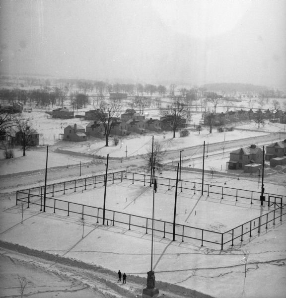 Elevated view of village with snow on the ground. A tennis court, flag pole, and pedestrians are visible in the foreground. Behind the tennis court are roads and dwellings.
