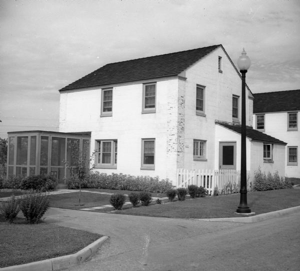 Exterior of two-story house with brick work on front corner. There is an enclosed patio extending from the house towards the front drive. A street lamp is on the right of driveway, and shrubbery lines the base of the house, as well as in the front yard.