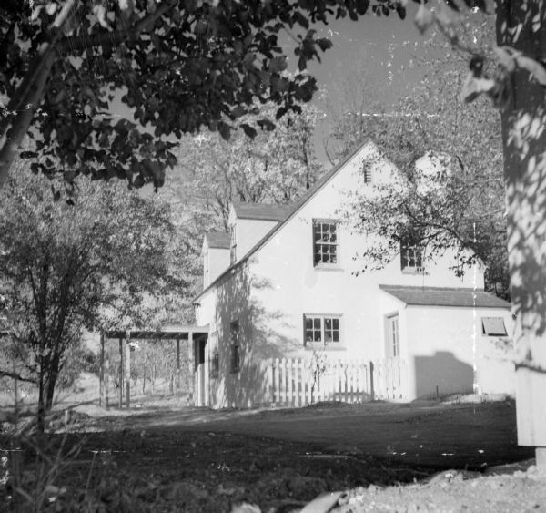 Exterior of a freshly built two-story house located at 293 East Orchard Court. The house has a covered patio. There is no driveway in place yet, and a freshly planted sapling is visible. Larger trees surround the house.