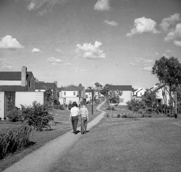 Two people, most likely a woman or girl, and a boy, are walking on a footpath with houses and young trees on either side of the path.