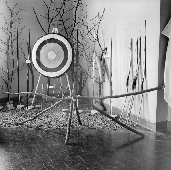 Display of bows, arrows and a target in a corner with tree branches and gravel at a hobby show. There are labels on some of the items.