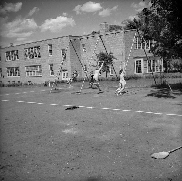 Four children, one boy and three girls, playing on a a swing set outside of a brick building, probably a school.