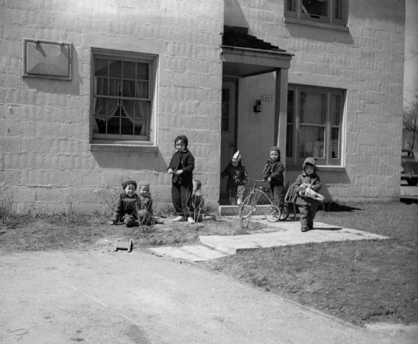 Seven children wearing coats and hats play in front of a roofed entrance of two-story house. One child has a tricycle, another is holding a doll. An automobile can be seen in the background.