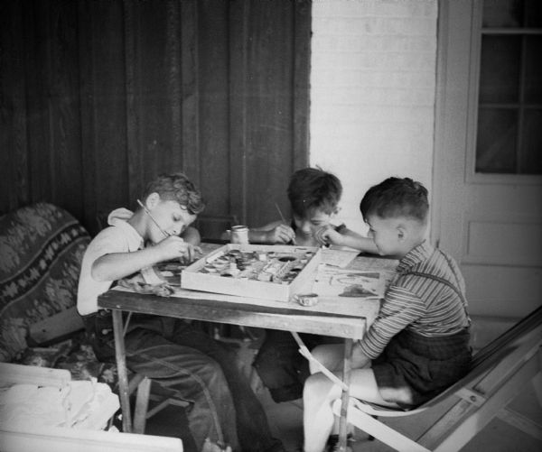 Three boys painting with watercolors at a folding table. They appear to be on a porch.