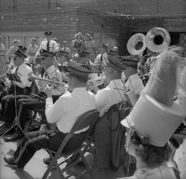 Band, in full regalia, playing tubas, clarinets and trumpets as well as other instruments, outside at a Fourth of July parade. There is a brick building in the background.